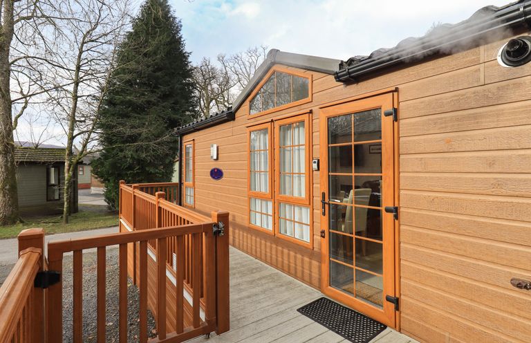 The exterior of a wooden cabin with a deck and railing in a wooded area at Squirrels Snug in Pony Meadow 1 White Cross Bay near Windermere