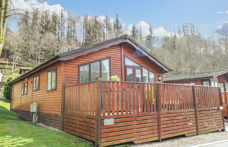 A wooden lodge with a fenced deck surrounded by grass and trees at Limefitt Lodge Fellside 8 Limefitt Park near Windermere
