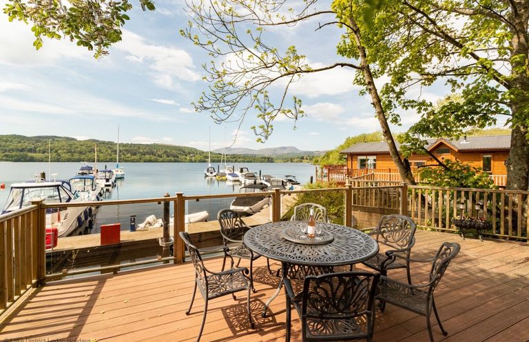 A wooden deck with a metal table and chairs overlooking a lake with boats and a wooden cabin nearby at Waterbird Lakeview 4 White Cross Bay near Windermere