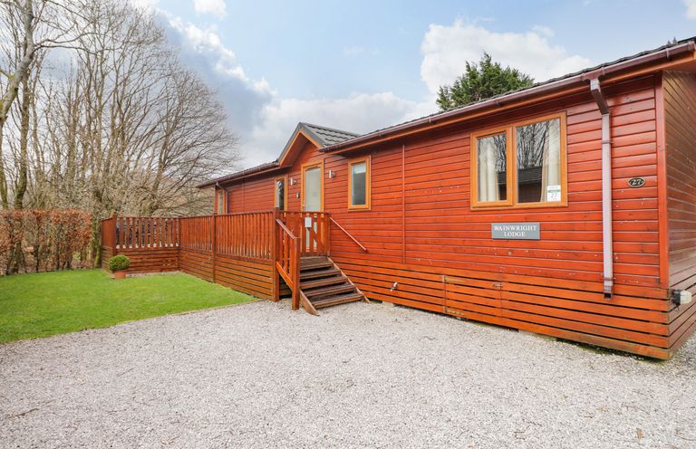 A wooden lodge with a small porch and steps on a gravel area next to green grass and trees at Wainwright Lodge Kirkstone 27 Limefitt Park near Windermere