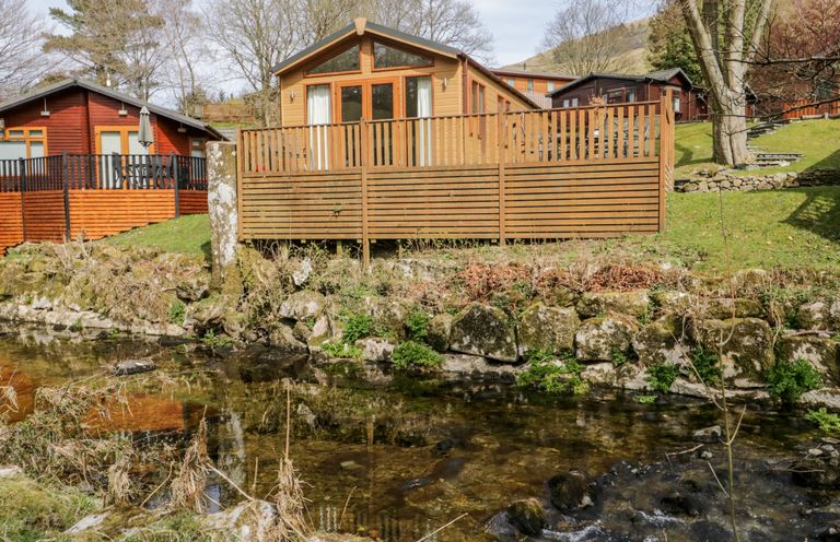 A wooden cabin with a deck next to a stream in a grassy area with trees at Troutbeck Lodge Beckside 6 Limefitt Park near Windermere