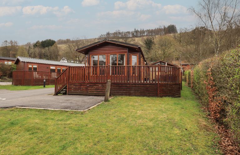 A wooden cabin with a fenced deck in a grassy area near trees and hills at Striding Edge Lodge Ullswater 9 Limefitt Park near Windermere