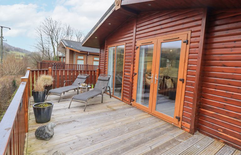 An outdoor wooden deck with two lounge chairs and potted plants outside a wooden cabin with glass doors at Horseshoe Lodge Wansfell 5 Limefitt Park near Windermere