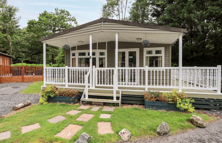 A front view of a small cabin with a covered porch and white railing surrounded by grass and trees at Pinehaven Lodge in Grasmere 6 White Cross Bay near Windermere
