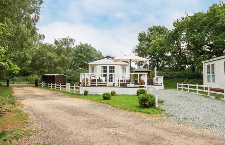 A small light-colored house with a deck and outdoor furniture next to a gravel driveway with trees in the background at Lakeside View in Pentney near King's Lynn