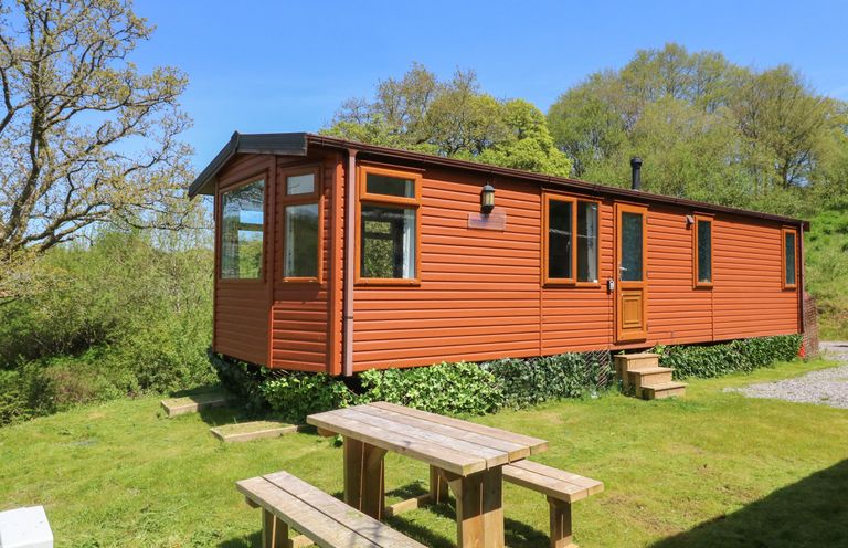 A wooden lodge with windows and a door next to a wooden picnic table and benches on grass at Valley Lodge in Bampton Devon