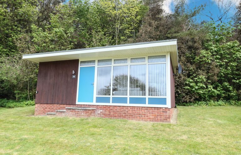 A small building with a blue door and large windows surrounded by grass and trees at Chalet 126a in Cromer