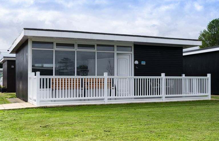 A black cabin with large windows a white door and a white fence on a grassy lawn at 57 Cherry Park in Chapel St Leonards