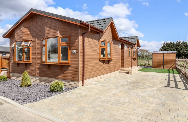 Exterior view of a brown wooden single-story house with multiple windows paved driveway and small garden with bushes at 12 Bucklesham Park in Bucklesham near Trimley St Martin