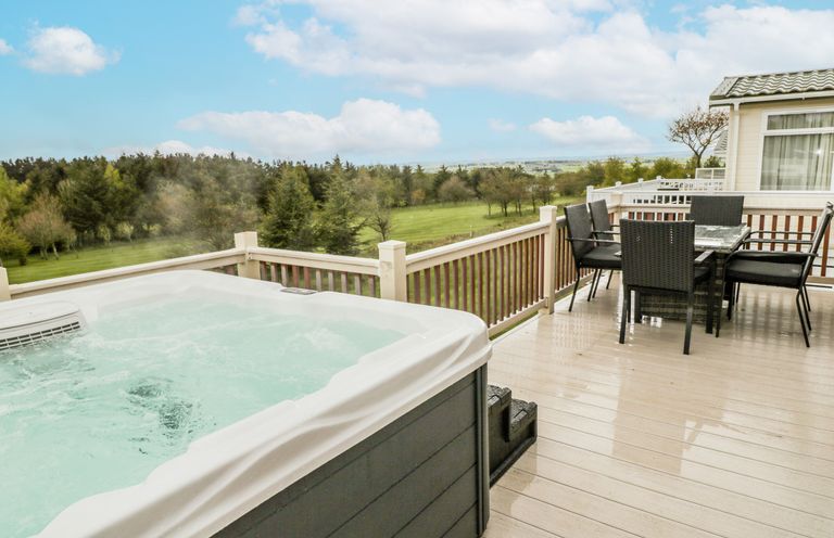 An outdoor deck with a hot tub and a dining table with chairs overlooking a grassy and tree filled landscape at Woodland View Lodge Swarland near Felton