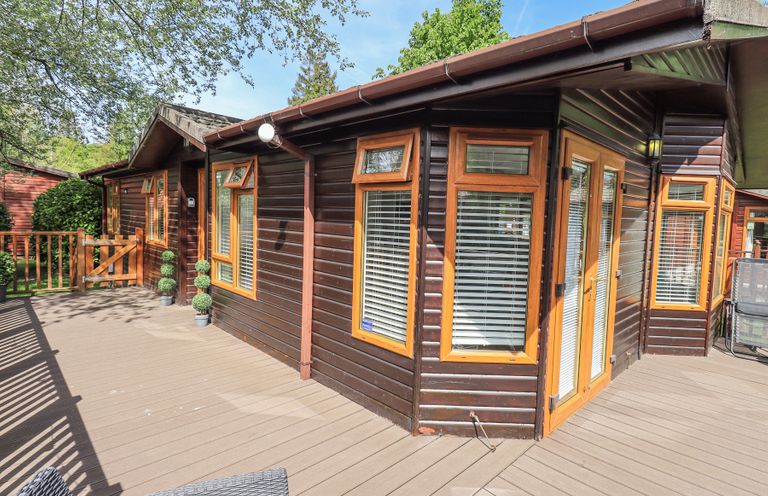 An outdoor wooden deck area with a dark brown wooden cabin with multiple windows and a door at Grizedale Lodge Langdale 18 White Cross Bay near Windermere