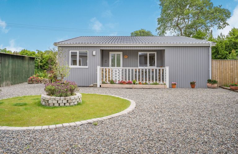A single story gray house with a small porch and flower beds in front at Caldey Lodge (Bier Wood Lodges) in Manorbier