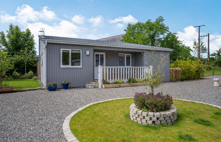 A gray single-story house with a small porch and gravel driveway with a circular planted area in front at Ramsey Lodge Bier Wood Lodges in Manorbier