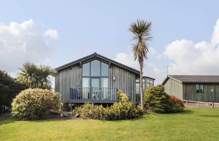 A wooden exterior house with large windows and a balcony surrounded by palm trees and bushes at Sea View Lodge Eastfield Hall near Warkworth