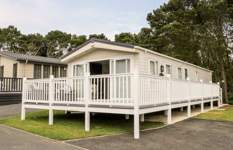 A beige mobile home with a white railing deck surrounded by trees at The Rodley Pines in Cayton