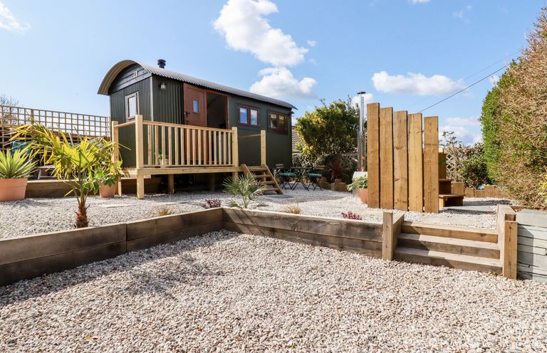 An outdoor area with a small wooden cabin on stilts a raised wooden platform a gravel ground and wooden steps at Badgers Rest in Tintagel