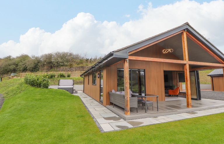An exterior view of a wooden house with a covered patio and outdoor seating on a lawn at Pool View in Meifod Powys