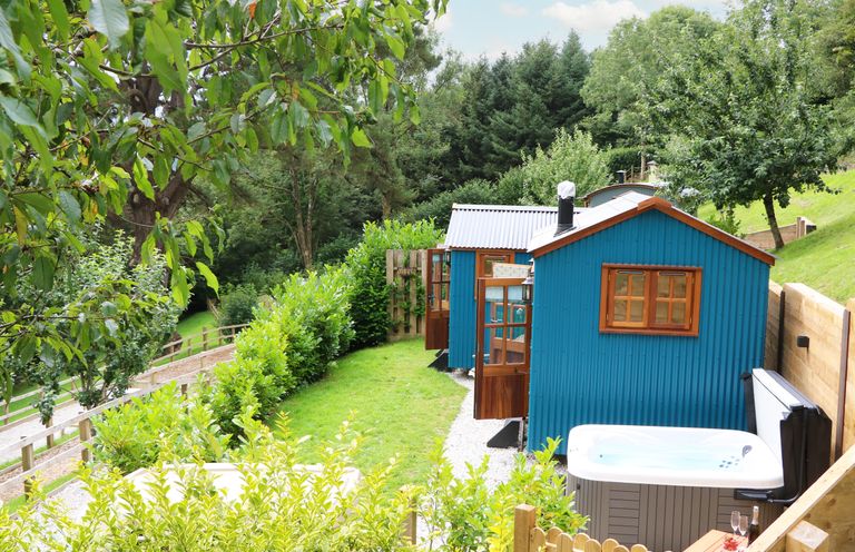 Two blue tiny houses with wooden doors and window frames in a green garden with a hot tub on a hillside at Little Silver Pheasant in High Bickington