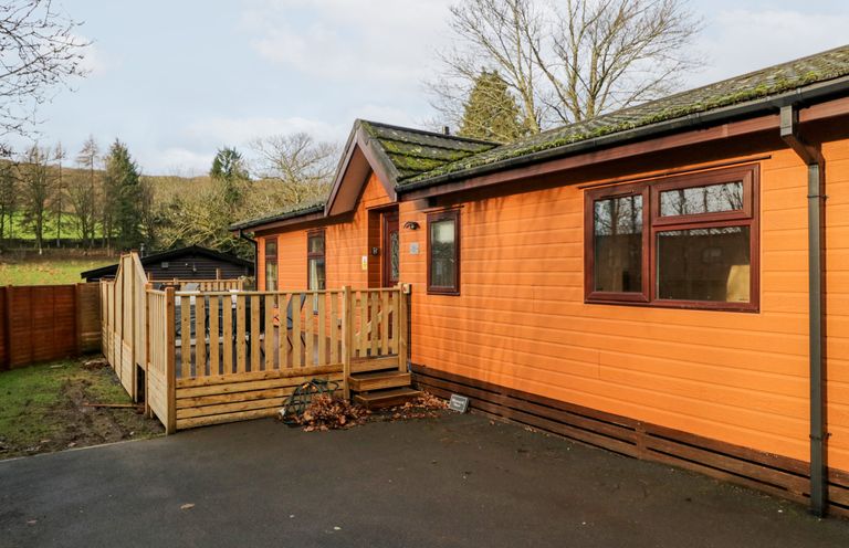 An orange wooden cabin with a fenced porch and trees in the background at Wansfell View in Limefitt Park near Troutbeck
