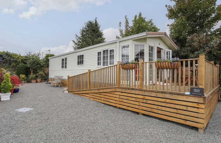 A mobile home with a wooden deck and flower baskets on gravel at Tegfryn Bach in Llangoed near Beaumaris