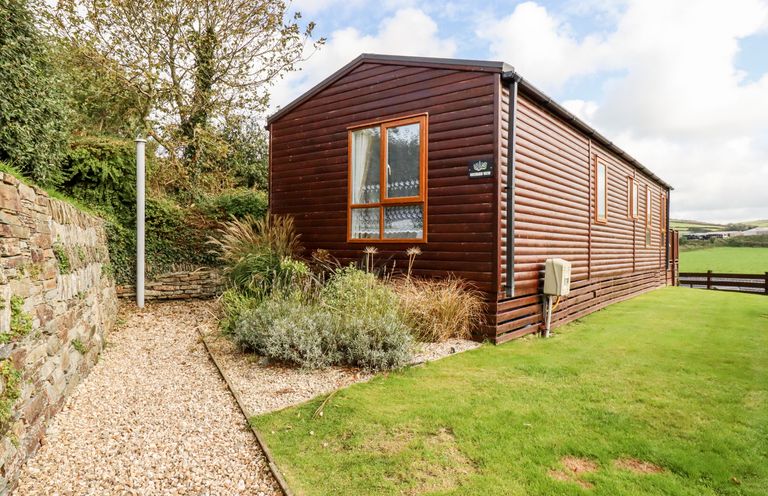 A brown wooden house with a window surrounded by grass and gravel path by a stone wall at Orchard View in Bossiney near Tintagel