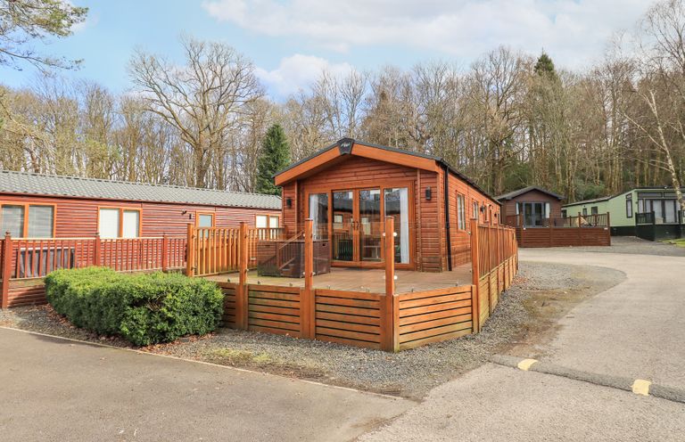A wooden lodge with glass doors and a fenced deck in a park with other lodges and trees at Windermere View Pony Meadow 37 White Cross Bay near Windermere