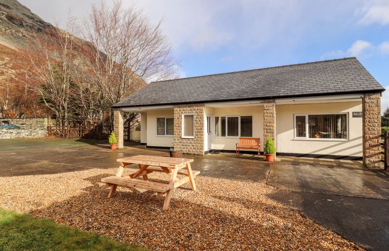 A single-story house with a patio bench and a wooden picnic table on gravel at Awel Y Berwyn in Llangynog