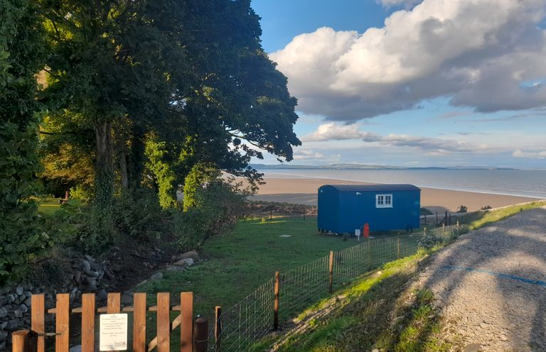 A blue shepherds hut near a sandy beach with trees and a wooden fence at Seashore Shepherds Hut @ Moat Farm in Aldingham near Ulverston