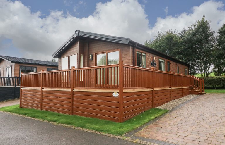 A brown wooden cabin with a surrounding deck and railing near a paved driveway and grass at The Warren in Hellifield