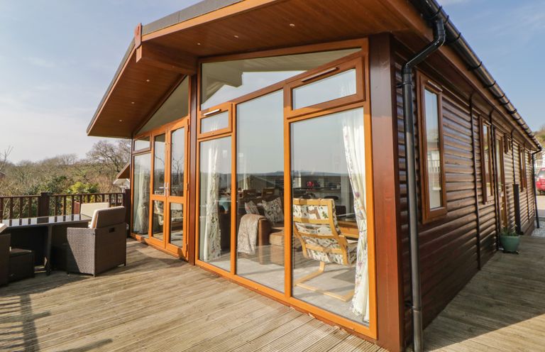 A wooden cabin with large glass windows and a deck with outdoor furniture at Meadow Lodge in Newton Ferrers