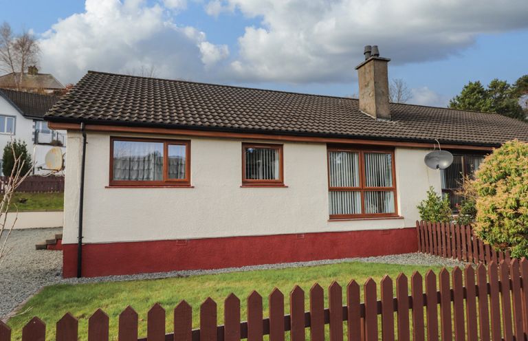 The exterior of a single-story house with a brown tiled roof and white walls with red trim enclosed by a wooden fence at The Hideout in Portree