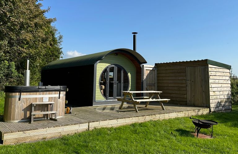 A wooden pod with curved roof next to a wooden hot tub and shed on a wooden deck with a picnic table and a fire pit on grass at Lime Kiln in Blagdon