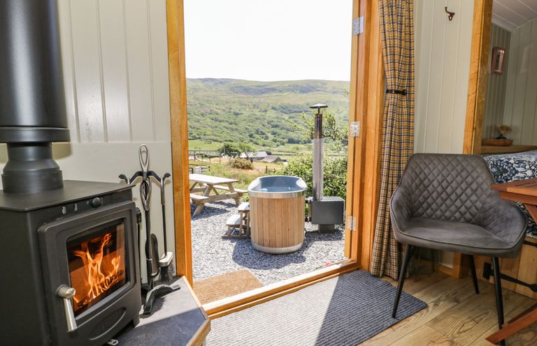 An interior view of a room with a wood burning stove a chair and an open door leading to an outdoor wooden hot tub and picnic table with hills in the background at Llethr - Shepherds Hut in Llanbedr