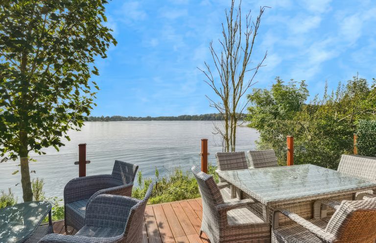 An outdoor deck with wicker chairs and glass tables overlooking a lake with trees at Gerrida Lodge South Lakeland Leisure Village near Tewitfield