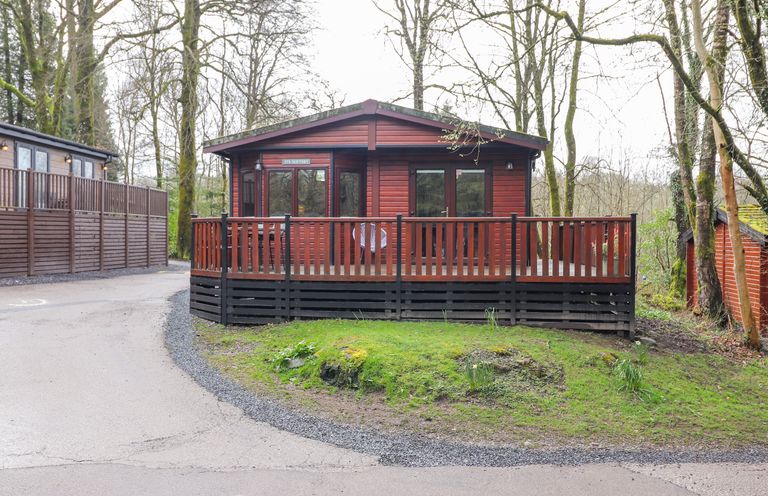 A wooden cabin with a fenced porch surrounded by trees at Alfresco Lodge in Skiptory Howe 37B White Cross Bay near Windermere