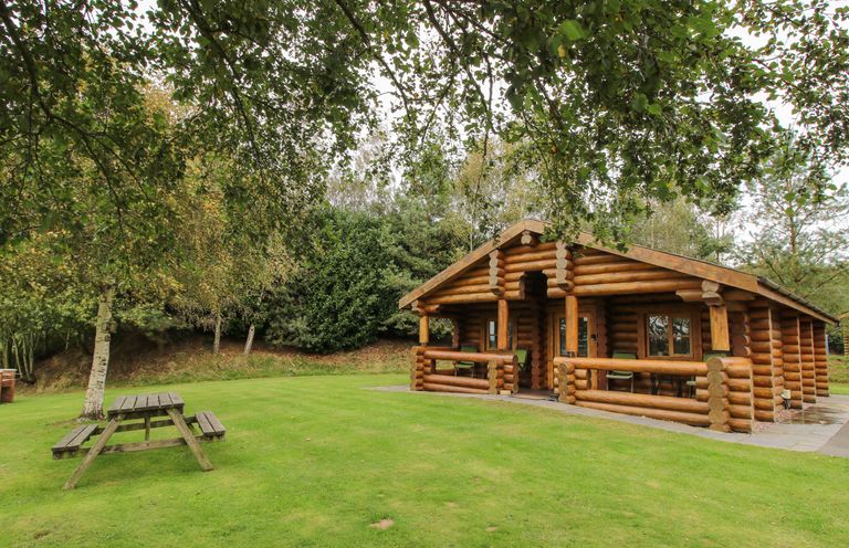 A wooden log cabin with a porch and green chairs on a lawn with a picnic table and trees at Brynallt Country Park Frankton near Ellesmere