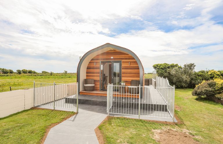 An arched wooden cabin with two chairs on the porch and a fenced patio in a grassy area at Caban Cariad in Bryngwran near Bodedern