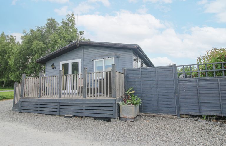 A gray wooden cabin with a fenced porch and plants outside at Wayside Lodge in Aston On Clun