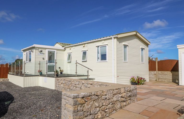 A cream-colored mobile home with a glass railing and stone wall outside at The Lodge in Rhosybol
