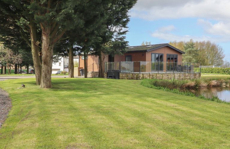A wooden house with a glass railing on a stone base near a body of water and trees on a grassy lawn at Ruperts Retreat in Nateby near Garstang