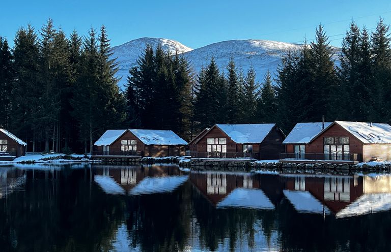 Snow covered cabins by a lake with pine trees and mountains in the background at Ptarmigan Lodge in Glengoulandie near Aberfeldy