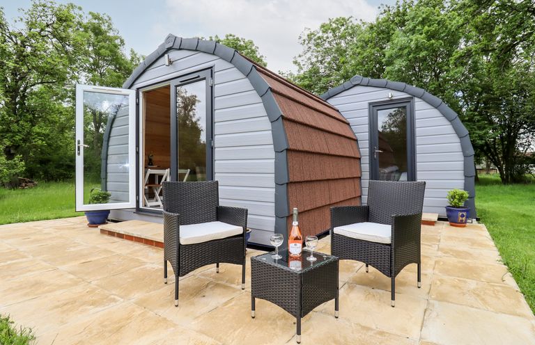 Two small pod-shaped cabins on a patio with two chairs and a table holding a bottle and two glasses at Meadow Lodge Berrys Place Farm in Churcham