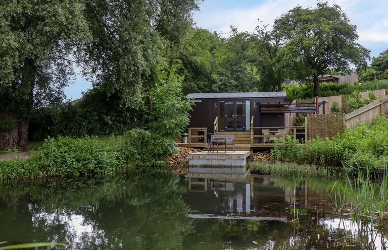 A small cabin with outdoor seating overlooking a pond surrounded by trees and greenery at The Shepherds Hut at Bridge Lake Farm & Fishery in Chacombe