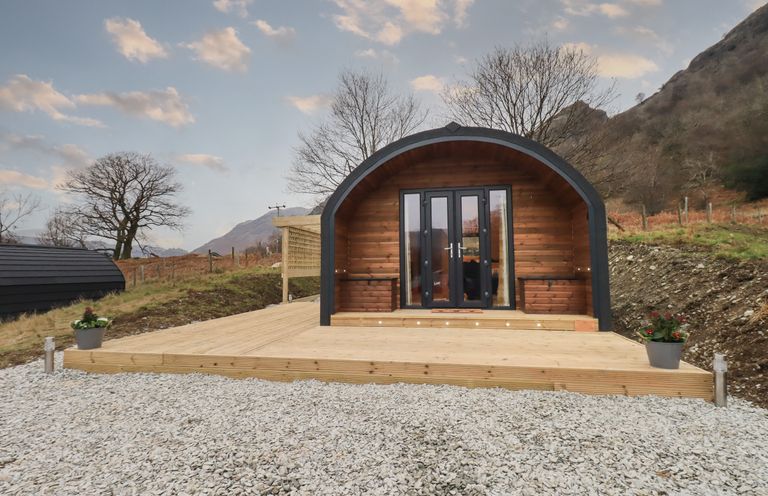 A wooden pod with glass double doors on a deck surrounded by gravel and trees at The Stag - Crossgate Luxury Glamping in Hartsop near Glenridding