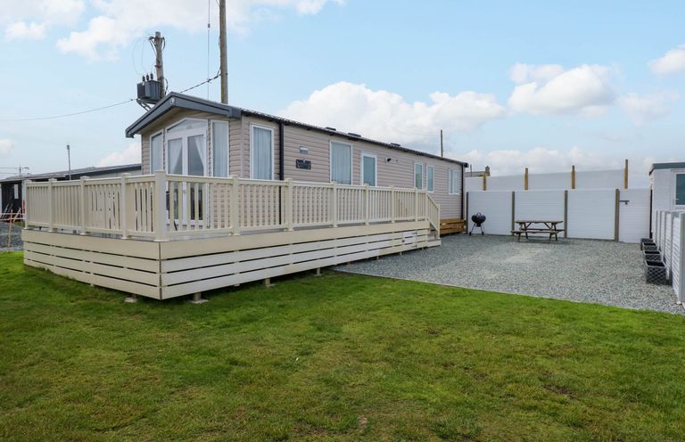 A mobile home with a surrounding deck and fenced gravel area with picnic table and barbecue on grass at Trewan Lodge in Bryngwran near Rhosneigr