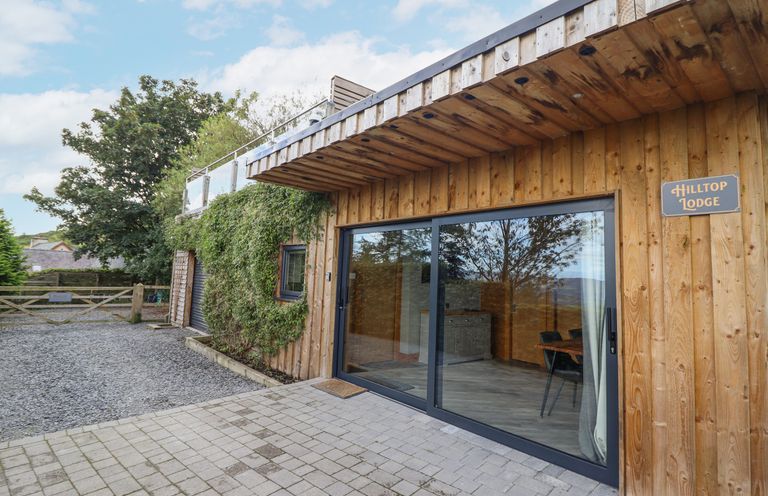 The exterior of a wooden building with large sliding glass doors and a gravel driveway at Hilltop Lodge in Corwen