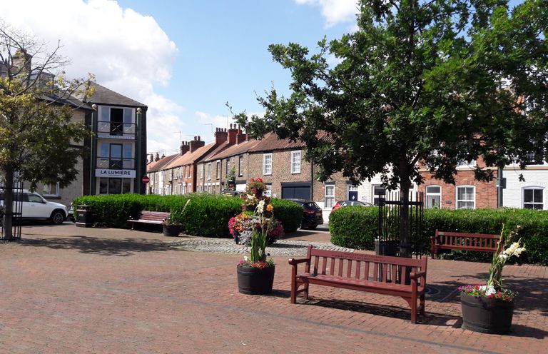 A small urban park with benches flower pots a tree and residential buildings in the background at No.37 in Beverley