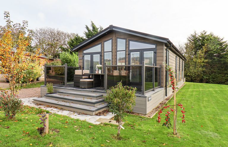 A cabin with a wooden deck and outdoor seating in a garden with small trees and grass at Hideaway Lodge in Runswick Bay