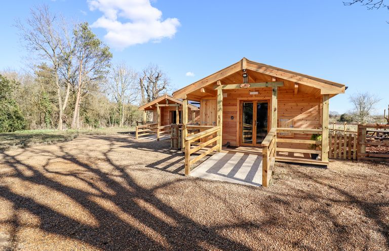 Two wooden cabins with porches on a gravel area surrounded by trees at Clover Lodge in Theberton near Leiston