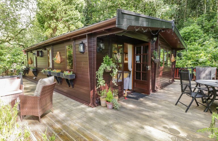 An outdoor wooden deck with chairs and tables around a brown wooden cabin surrounded by trees at Scafell Lodge Skiptory Howe 7 White Cross Bay Holiday Park near Windermere